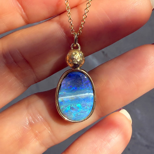 An Australian Opal Necklace by NIXIN Jewelry is shown in a close-up image against a hand, emphasizing its meticulous details. This perspective provides an intimate look at the intricately hand-textured gold moon, featuring its flush-set diamond, and the mesmerizing natural blue boulder opal within its precise gold bezel. This detailed shot underscores the high jewelry craftsmanship and jewelry art of this rare opal and diamond necklace.