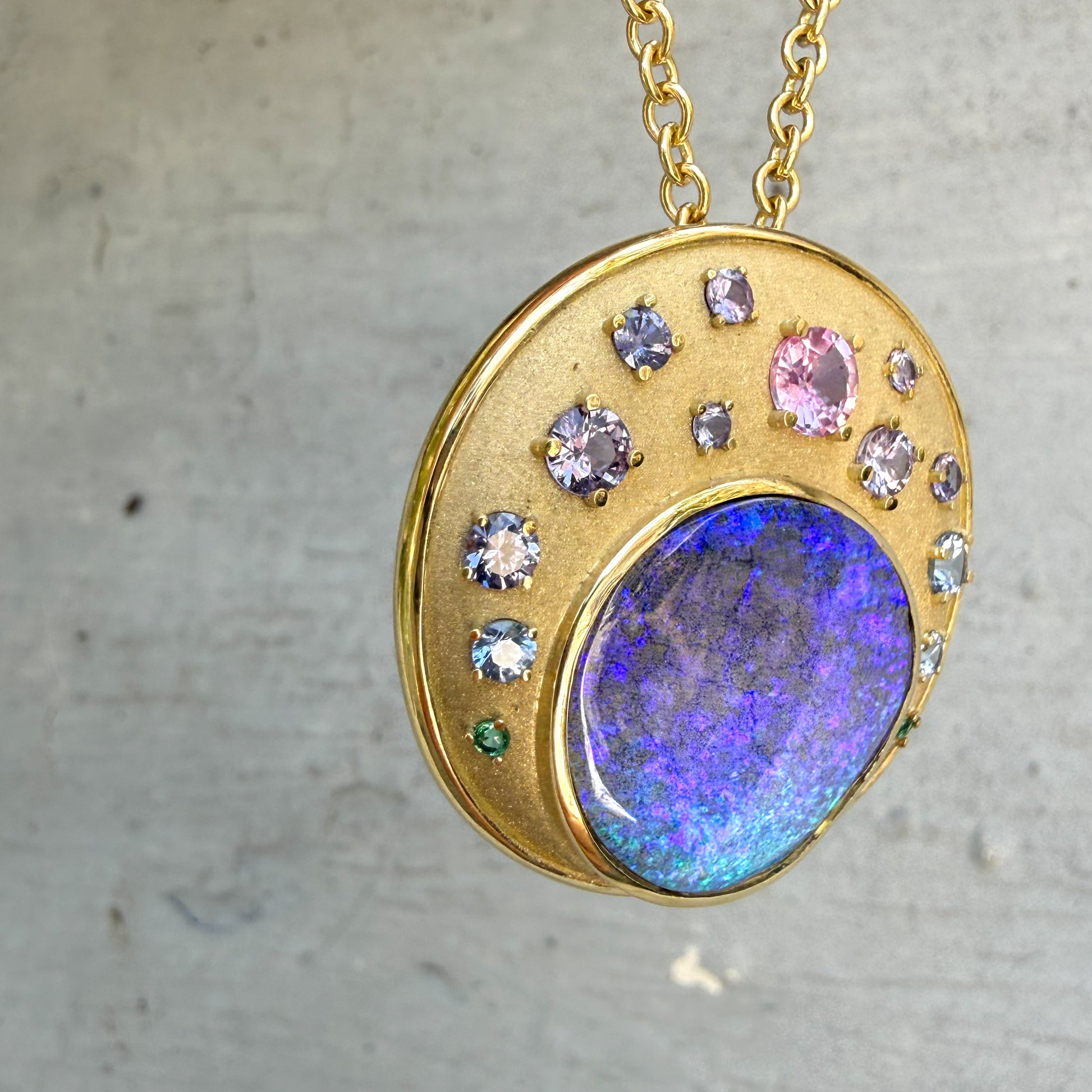 This close-up angle of an Australian Opal Pendant by NIXIN Jewelry emphasizes the play of color in the rare jewelry, from the deep blue and purple opal to the ombre sapphires and emeralds. The gold opal necklace features a rare opal and an artful combination of precious stones, all in a unique bezel and prong setting. This piece is a beautiful example of luxury jewelry art.