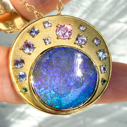 This close-up view features an Australian Opal Pendant by NIXIN Jewelry, showcasing a vibrant boulder opal in a secure bezel setting. The unique fine jewelry is adorned with an ombre array of pink, purple and blue sapphires, all held by gold prongs. The matte gold finish is evident in this natural daylight shot, highlighting the rare jewelry.