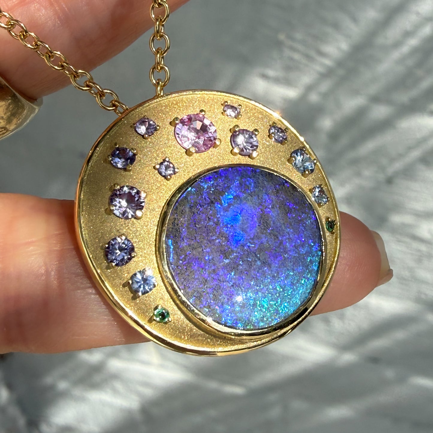 An Australian Opal Pendant by NIXIN Jewelry is displayed in this close-up, illustrating the stunning color play of the blue opal and purple sapphires. This unique fine jewelry is a true example of jewelry art, combining precious opal with an ombre-style sapphire arrangement. The gold opal necklace is clearly shown in this detailed view of the luxury jewelry.