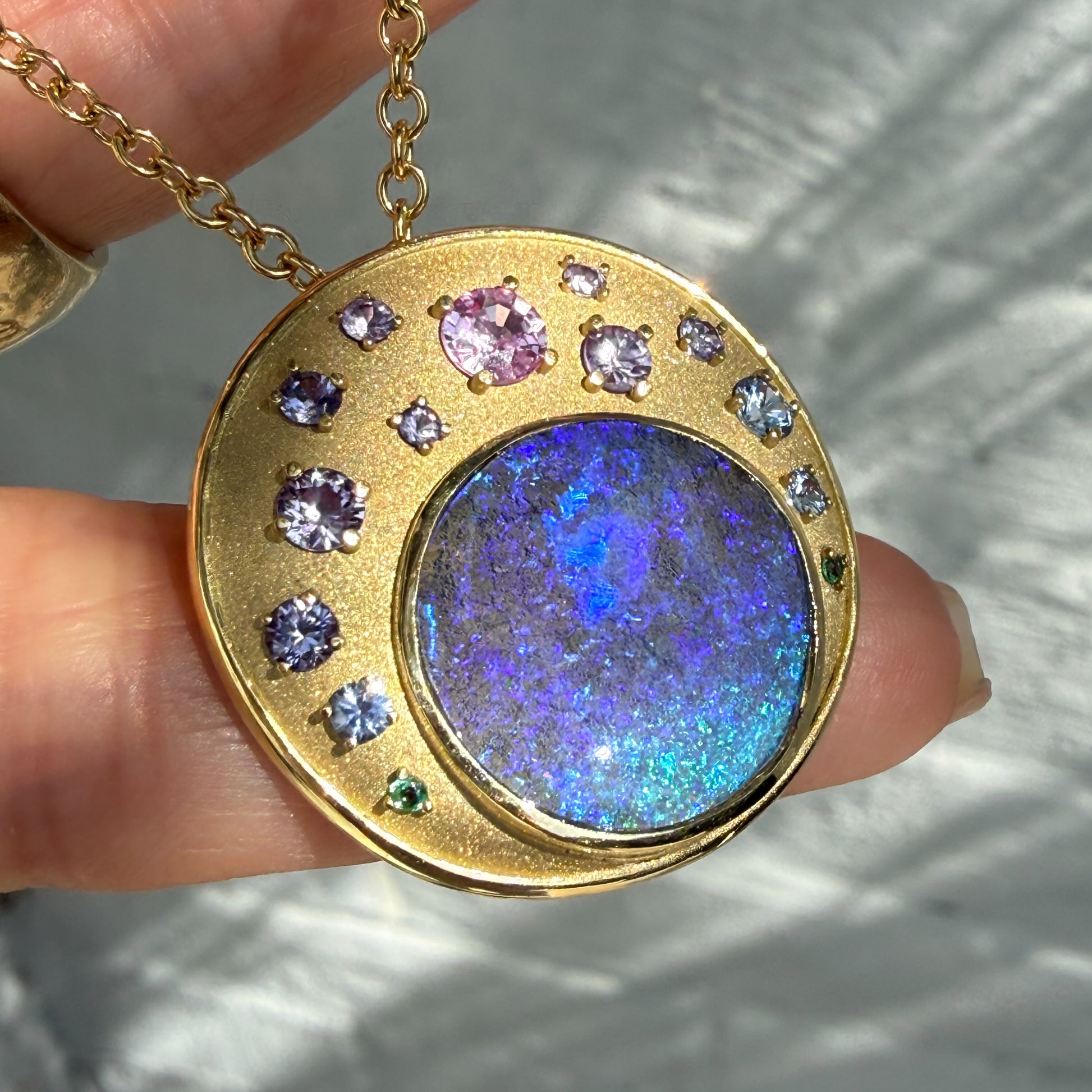 An Australian Opal Pendant by NIXIN Jewelry is displayed in this close-up, illustrating the stunning color play of the blue opal and purple sapphires. This unique fine jewelry is a true example of jewelry art, combining precious opal with an ombre-style sapphire arrangement. The gold opal necklace is clearly shown in this detailed view of the luxury jewelry.