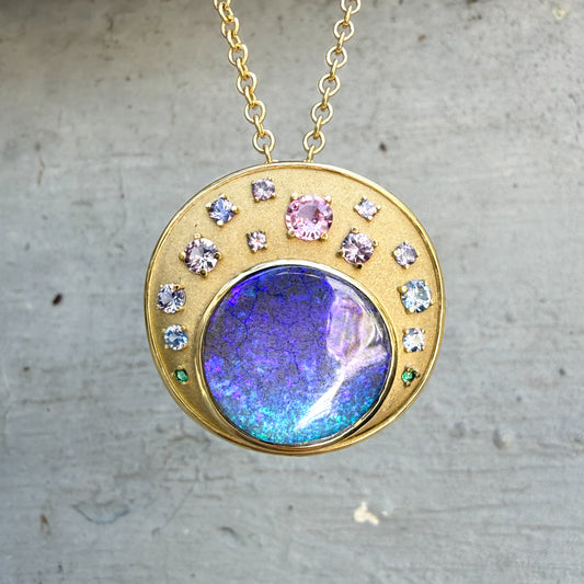This detailed photo shows an Australian Opal Pendant by NIXIN Jewelry, highlighting the boulder opal necklace against a neutral, concrete background. The piece's unique design features a natural opal and a vibrant ombre sapphires, set in warm, 18 gold. This unique fine jewelry is a perfect example of a women's luxury necklace.