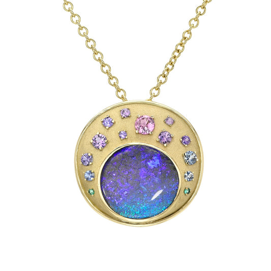 This professional studio shot of an Australian Opal Pendant by NIXIN Jewelry presents the rare jewelry against a clean white background. This unique fine jewelry piece features a boulder opal with a spectacular color play, surrounded by an ombre of natural sapphires, making it a truly unique opal necklace. The gold chain and sandblast finish of the women's luxury necklace are perfectly displayed.