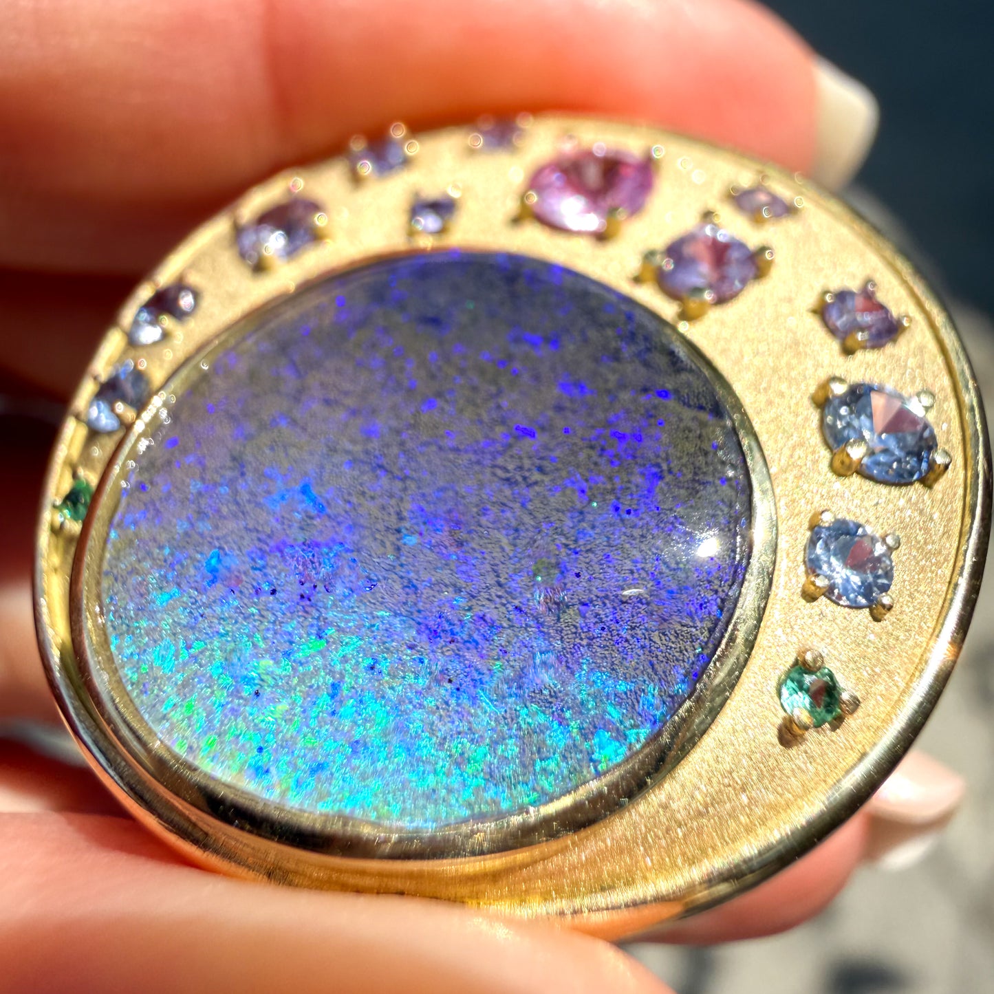 A magnified close-up photograph focused tightly on the natural opal of an Australian Opal Pendant by NIXIN Jewelry. The incredible texture of cracked, "parched earth" texture of the precious boulder opal stone is magnified, highlighting its rare jewelry quality. Only a few surrounding sapphires and the precise gold setting are visible, emphasizing the unique fine jewelry detail.