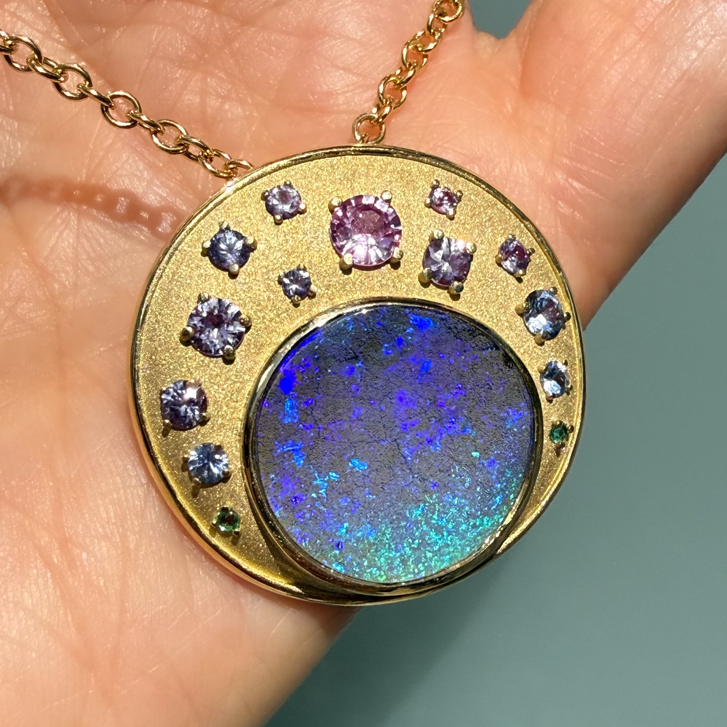 A clear shot showing the full front view of an Australian Opal Pendant by NIXIN Jewelry being held flat in a hand. The centerpiece is a precious opal, specifically a vibrant blue boulder opal with stunning color play, secured in a smooth bezel setting. Emeralds and an artful array of multi-colored sapphires in prong settings completes this unique opal necklace.