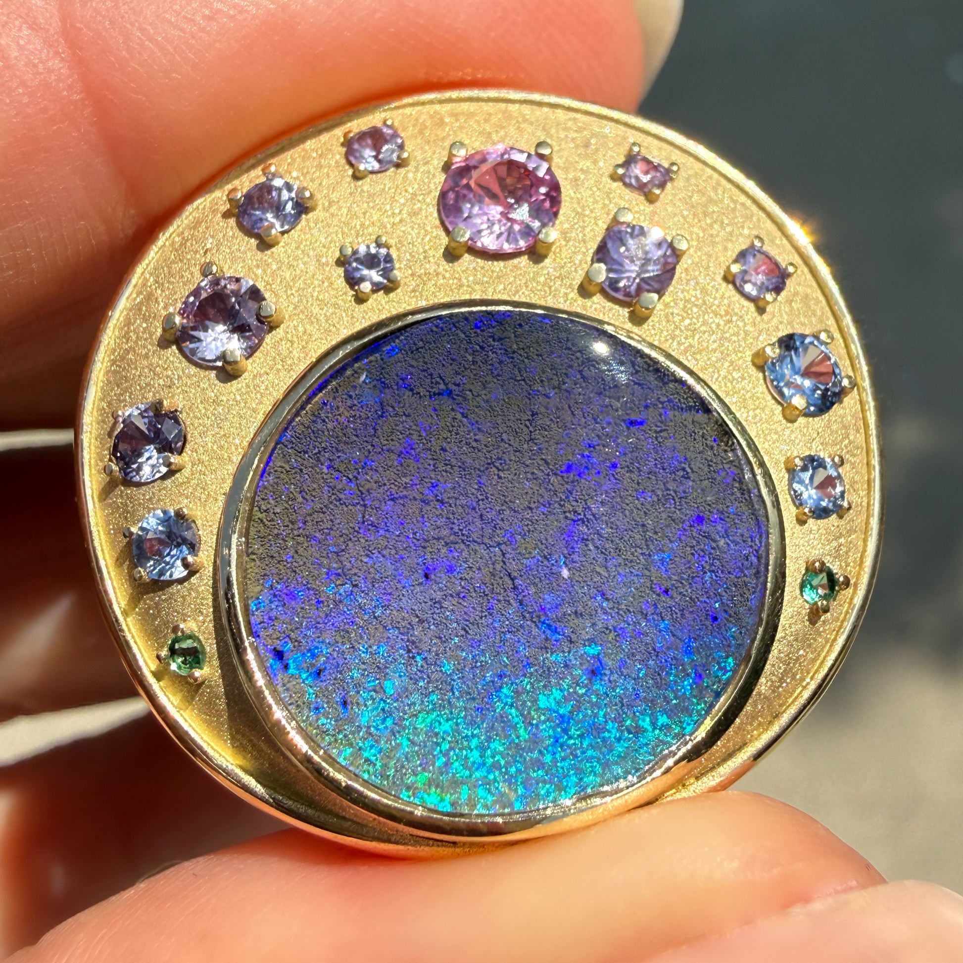 A detailed close-up shot showing the exceptional play-of-color within an Australian Opal Pendant by NIXIN Jewelry. The vibrant blue opal is held securely within a textured matte gold setting that features a meticulously matched ombre of multi-colored sapphires, punctuated by two vivid emeralds. This unique opal jewelry piece is a rare and luxurious choice for a boulder opal necklace.