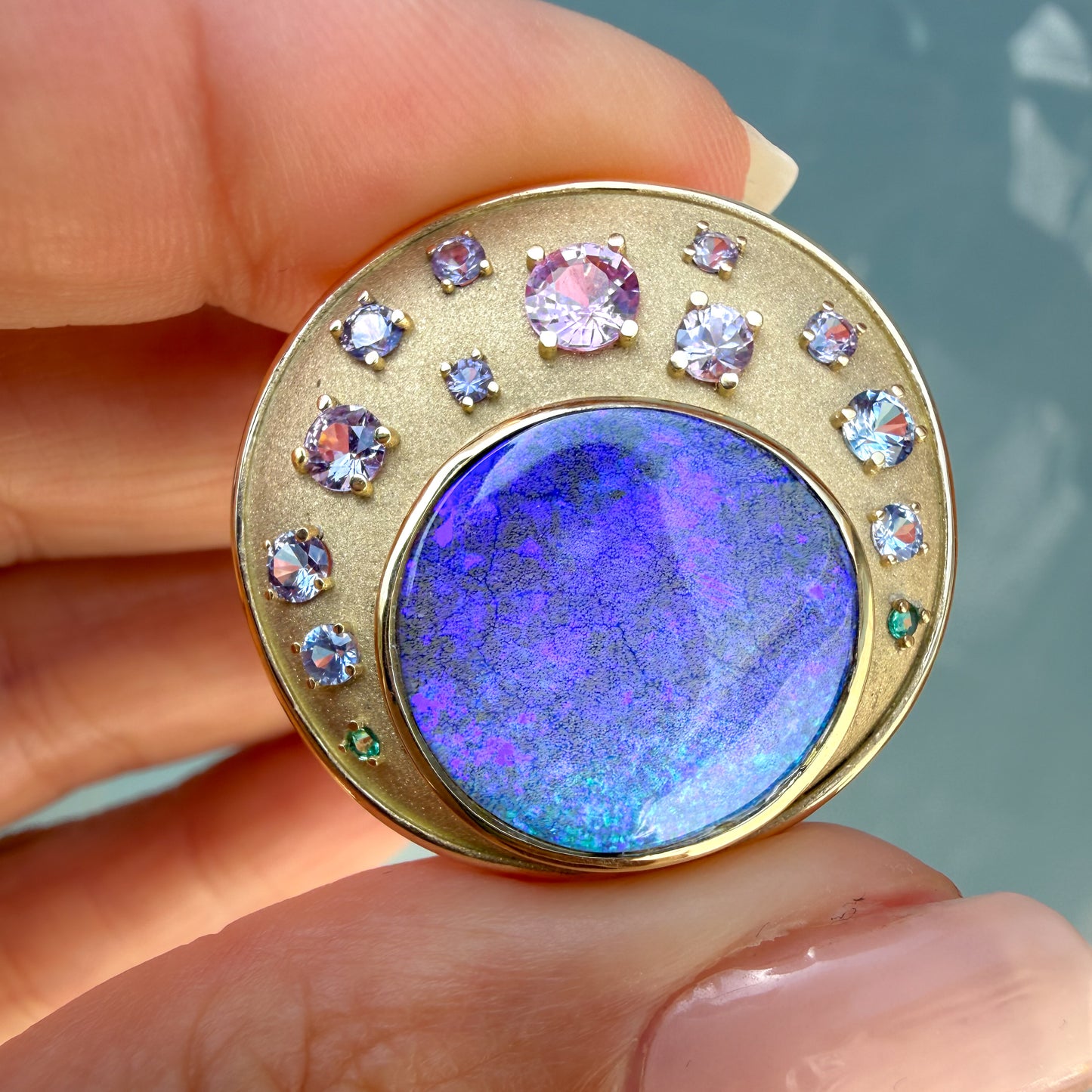 This extremely close-up photograph focuses tightly on the intricate details of an Australian Opal Pendant by NIXIN Jewelry. The large precious opal is set within a precise bezel setting and displays an intense saturated pattern with flashes of purple, blue and green. Small emeralds and sapphires in varying hues create a gradient around the natural opal, defining this unique opal necklace as jewelry art.