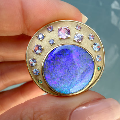 This extremely close-up photograph focuses tightly on the intricate details of an Australian Opal Pendant by NIXIN Jewelry. The large precious opal is set within a precise bezel setting and displays an intense saturated pattern with flashes of purple, blue and green. Small emeralds and sapphires in varying hues create a gradient around the natural opal, defining this unique opal necklace as jewelry art.