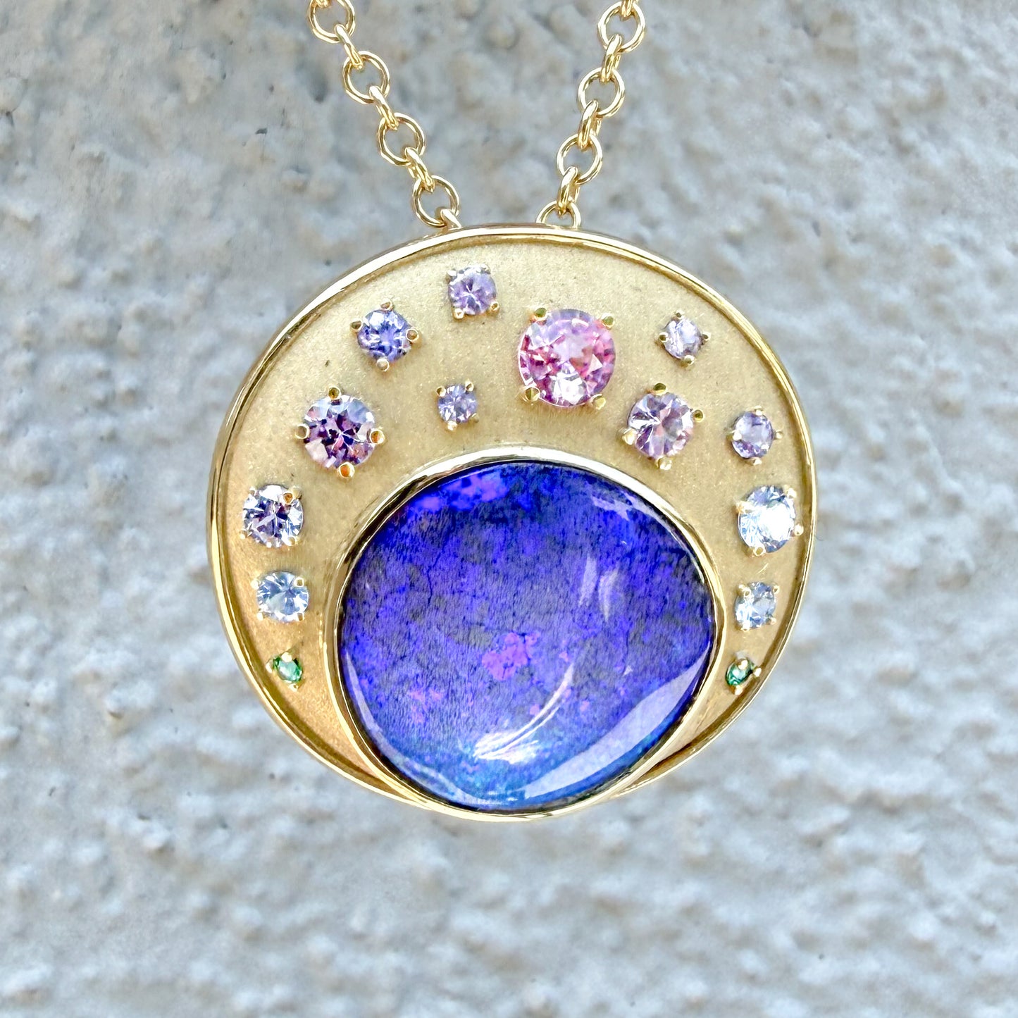 A clear, direct view of an Australian Opal Pendant by NIXIN Jewelry, with a grey textured stucco background providing visual contrast. The rich purple and blue opal is the centerpiece of this unique design, featuring a cascade of ombre sapphires and emeralds with a prominent pink sapphire. The gold opal necklace is a perfect blend of luxury and art, suitable for any collection.