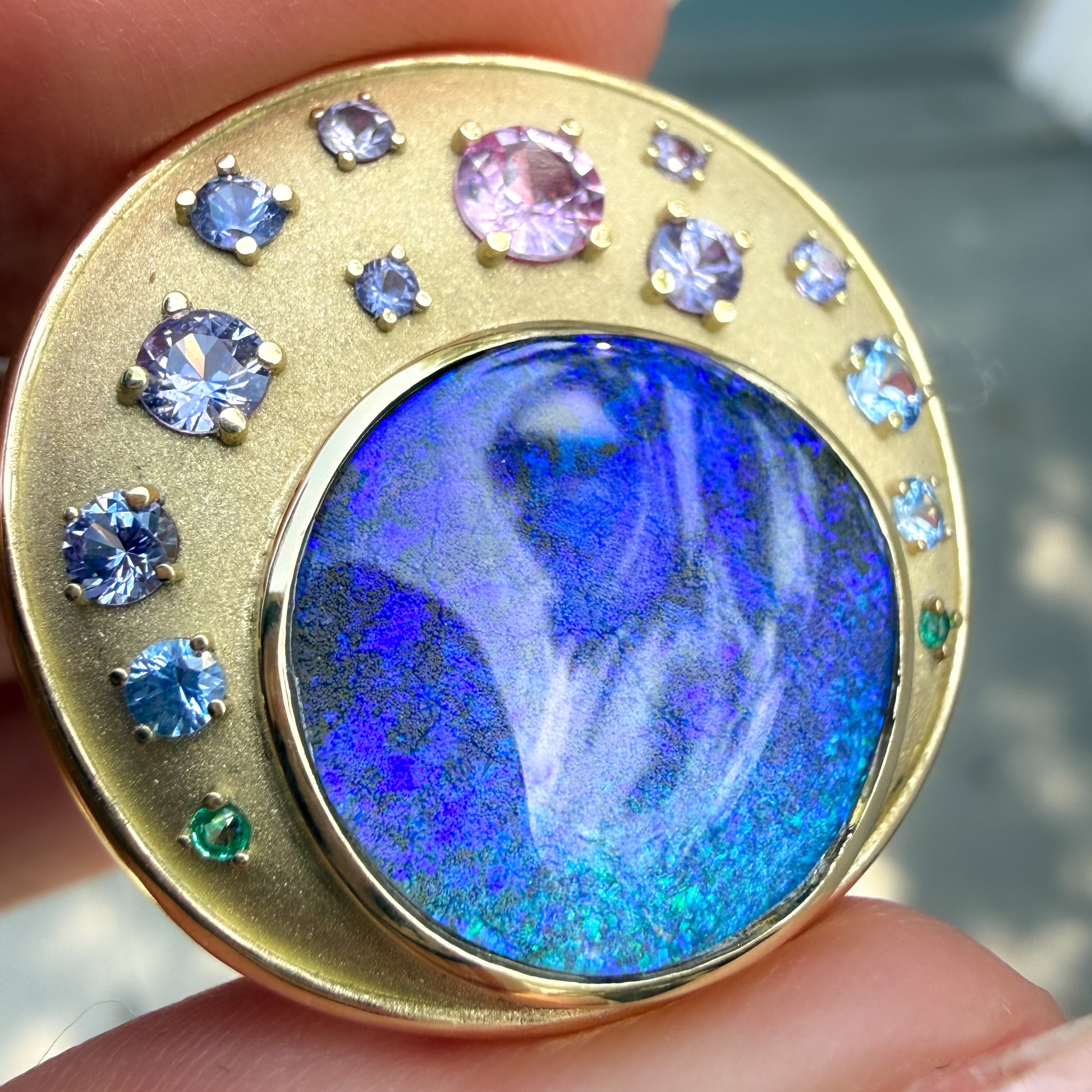 An Australian Opal Pendant by NIXIN Jewelry, carefully held between a finger and thumb for a detailed perspective on the prong and bezel settings. This unique fine jewelry piece showcases a vibrant natural opal with dramatic color play, surrounded by an ombre array of multi-colored sapphires in a secure setting. The textured satin finish of the 18k gold highlights the craft as wearable art.