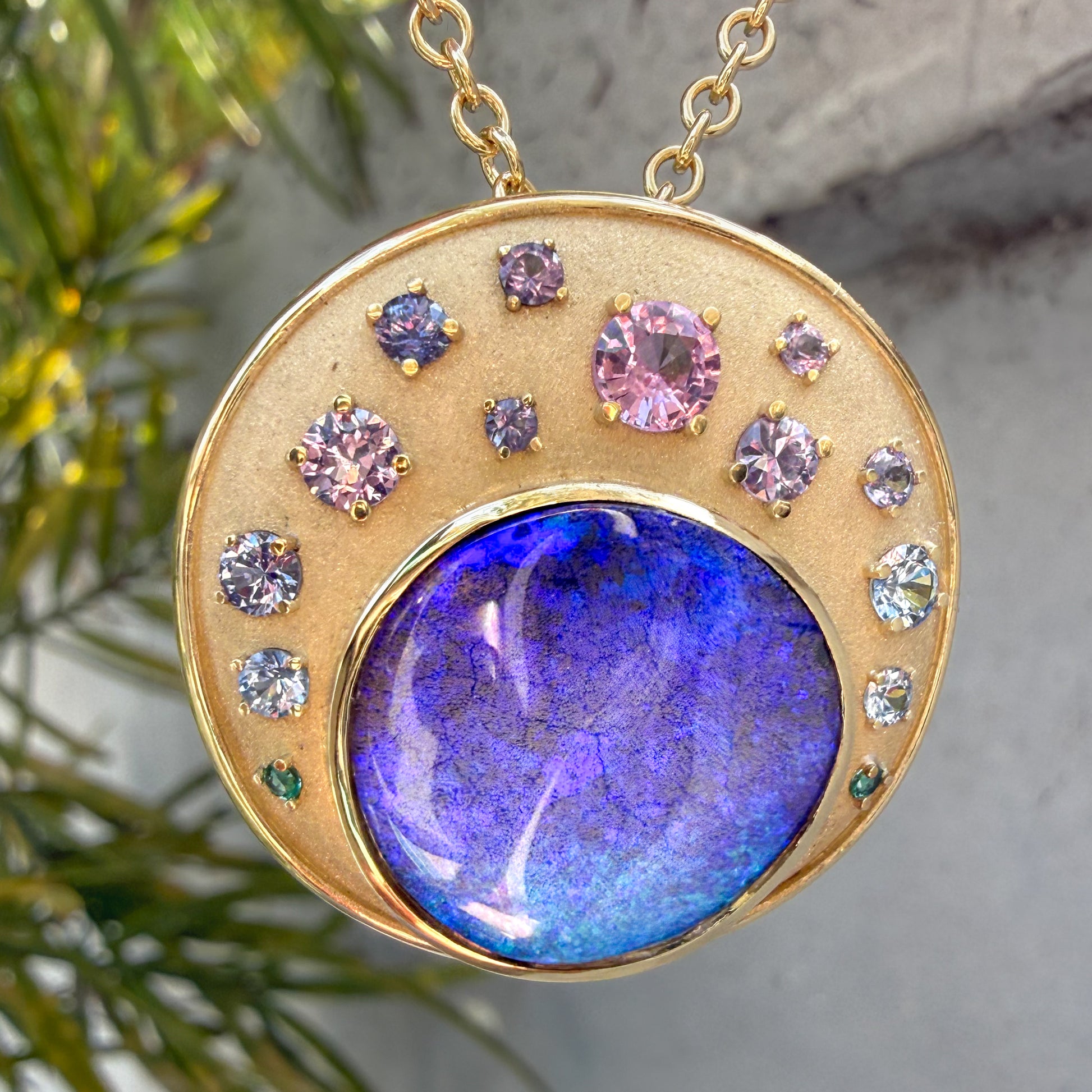 A close-up view of an Australian Opal Pendant by NIXIN Jewelry hanging from a delicate gold chain. The pendant features a large boulder opal with an ombre setting of precious sapphires against a blurred foliage background. This unique jewelry piece is a stunning example of nature-inspired luxury and art.