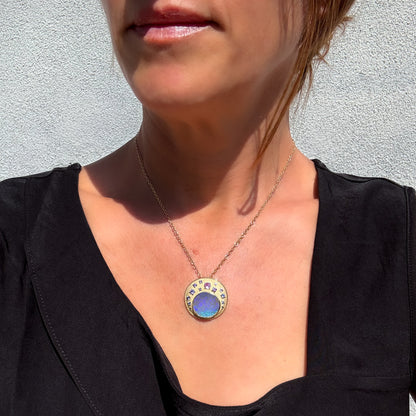 A woman wears an Australian Opal Pendant by NIXIN Jewelry, which showcases a boulder opal in a smooth bezel setting. This women's luxury necklace features an arrangement of small, multi-colored sapphires creating an ombre effect above the natural opal. The entire gold opal necklace has a beautiful matte gold texture. It's a true piece of jewelry art.