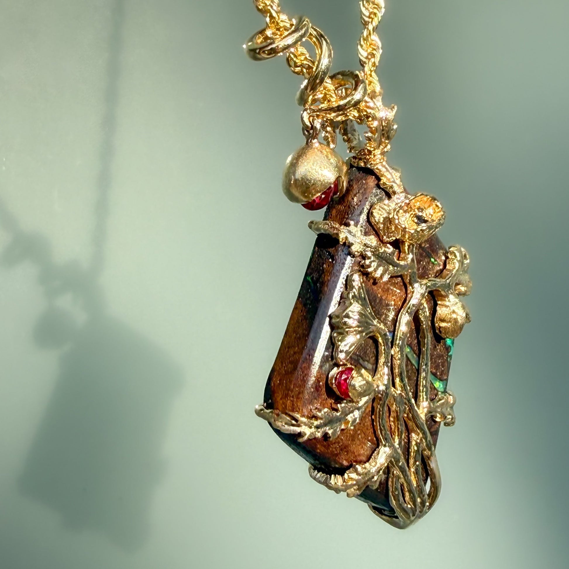 An Australian Opal Pendant by NIXIN Jewelry is captured at a three-quarter angle to showcase the dimensional 14k gold poppy vines as they emerge from the stone. These views highlight the jewelry art of the piece, where brilliant red rubies act as botanical accents against the vibrant green flashes of the boulder opal. This unique opal jewelry is a masterpiece of storytelling, blending luxury jewelry design with the raw beauty of natural opal.