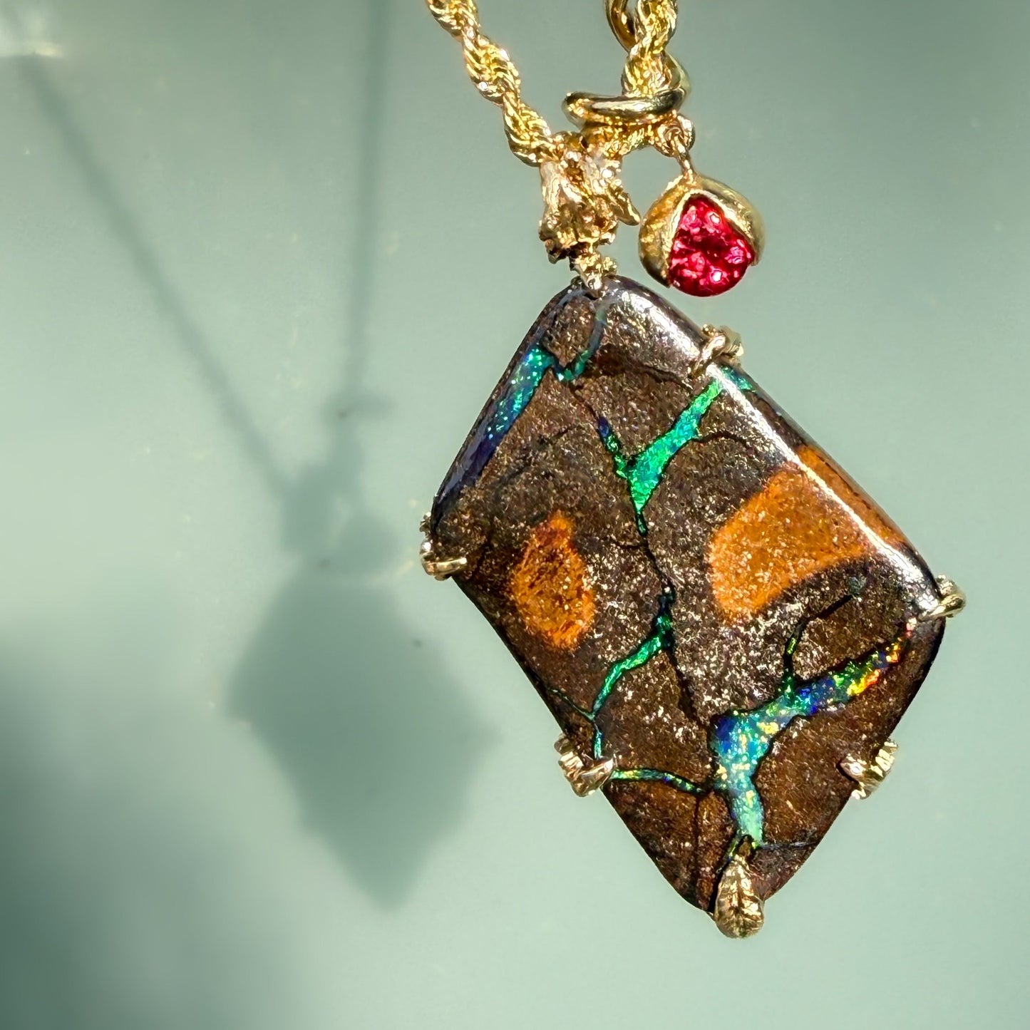 An Australian Opal Pendant by NIXIN Jewelry reveals a vibrant emerald-green leaf pattern with fiery red and cobalt blue sparkles within the natural opal stone. This rare opal jewelry is complimented by gold prongs and ruby accents, capturing the "Earth Remembers" theme through vivid jewelry art. It is a premier example of a gold opal necklace that appeals to collectors of unique opal jewelry.