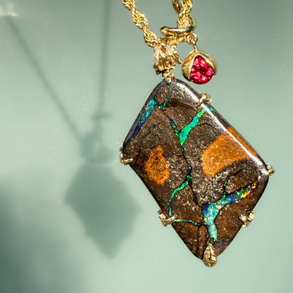 An Australian Opal Pendant by NIXIN Jewelry reveals a vibrant emerald-green leaf pattern with fiery red and cobalt blue sparkles within the natural opal stone. This rare opal jewelry is complimented by gold prongs and ruby accents, capturing the "Earth Remembers" theme through vivid jewelry art. It is a premier example of a gold opal necklace that appeals to collectors of unique opal jewelry.