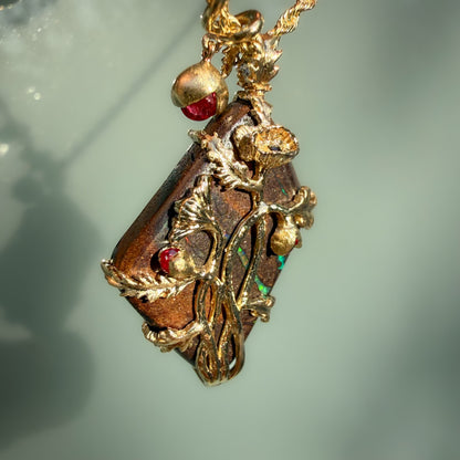 An Australian Opal Pendant by NIXIN Jewelry is presented in a dynamic, tilted profile to emphasize the high-relief 14k gold poppy blooms and organic vine textures. This luxury jewelry piece showcases how the natural opal and brilliant red rubies interact with light from a side-lit perspective, highlighting the depth of the artisan jewelry craftsmanship. It is an exquisite example of rare opal jewelry that combines sculptural jewelry art with high-end materials.