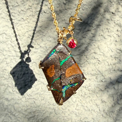 An Australian Opal Necklace by NIXIN Jewelry is presented with its shadow to illustrate the organic, botanical form of the boulder opal necklace. The interplay between the natural opal flashes and the brilliant red rubies showcases why this is considered rare opal jewelry. This unique opal necklace is a statement of resilience and beauty, embodying the pinnacle of jewelry art.