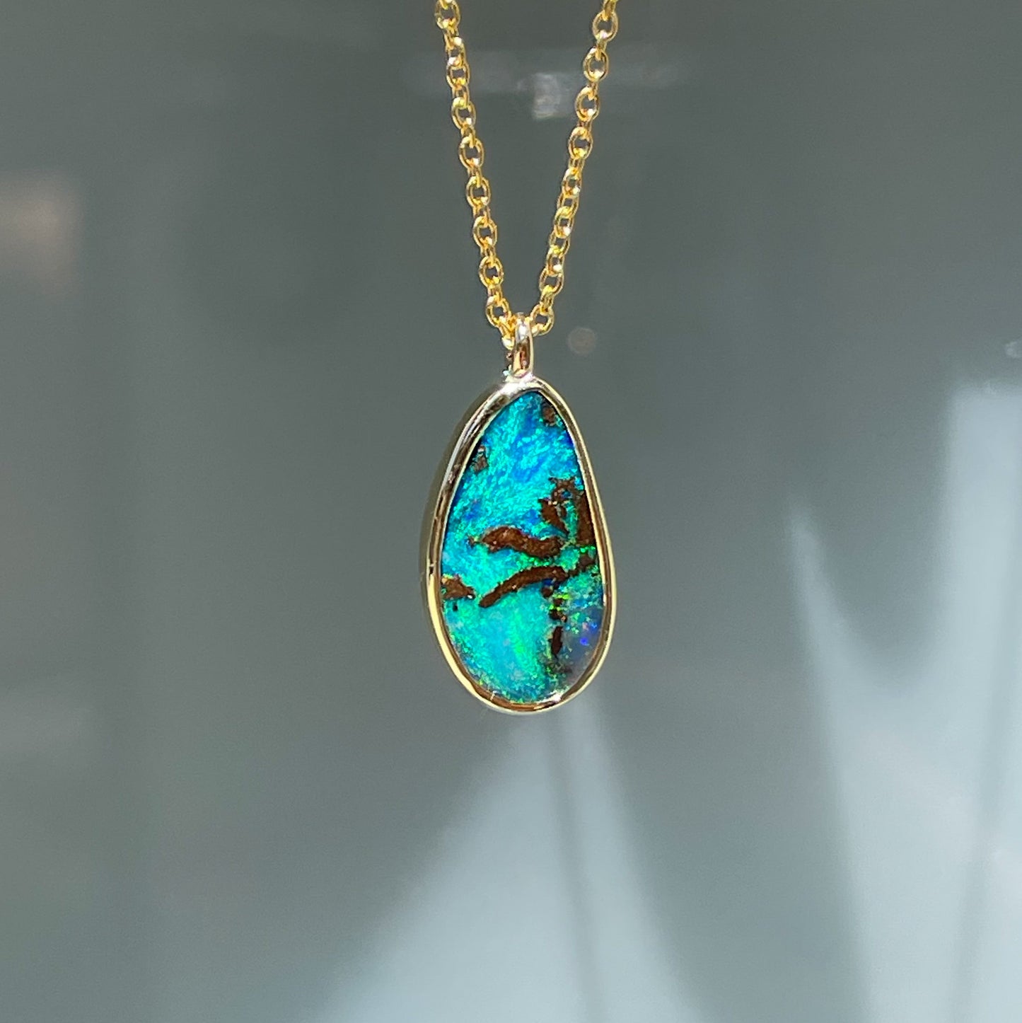 An Australian Opal Necklace by NIXIN Jewelry hanging in front of a frosted glass door. The natural opal is blue and green with inclusions that look like a palm tree. Made in 14k gold.