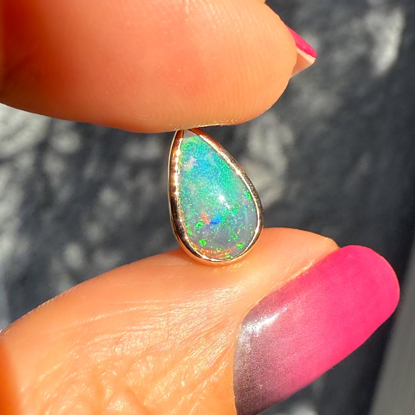 An Australian Opal Necklace by NIXIN Jewelry shown close-up, held between fingers to highlight the size and detail of the opal stone. This opal necklace features a striking bezel setting in 14K rose gold. The green and aqua colors make this ready-to-ship jewelry for opal pendant collectors.
