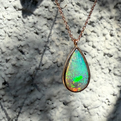 An Australian Opal Necklace by NIXIN Jewelry contrasted against a textured outdoor background to demonstrate light performance and versatility. The green fire of the crystal opal necklace shines brightly against the 14K rose gold opal necklace chain. This unique opal necklace represents the elegant craftsmanship and jewelry art.