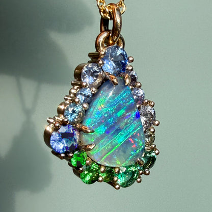 A vibrant front view of an Australian Opal Necklace by NIXIN Jewelry showcases a brilliant blue opal surrounded by a gradient of colorful gemstones. This unique opal jewelry features an ombre jewelry effect transitioning from deep blue sapphires to emeralds and bright green tsavorite garnets.