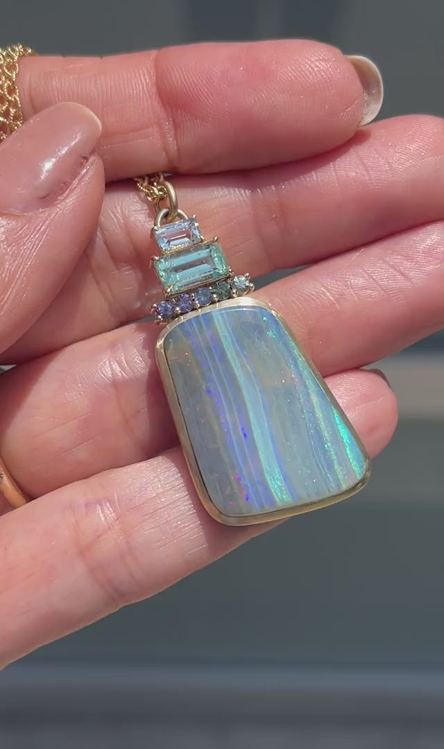 Video of an Australian Opal Necklace by NIXIN Jewelry held in sunlight. The gold opal necklace is made with a Boulder opal, ombre sapphires, an emerald and aquamarine.