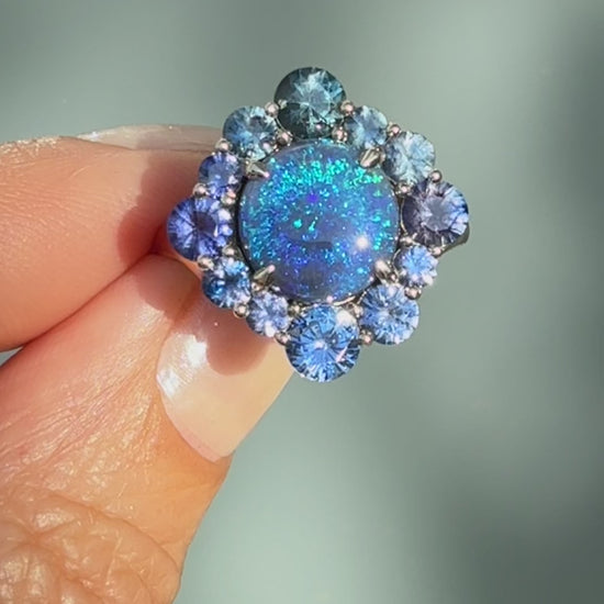 An Australian Opal Ring by NIXIN Jewelry. This video captures the close-up rotation of the magnificent black opal and sapphire opal ring cluster in its prong setting. The movement highlights the depth of color and the brilliance of this luxury ring, showcasing the meticulous craftsmanship of this unique jewelry piece.