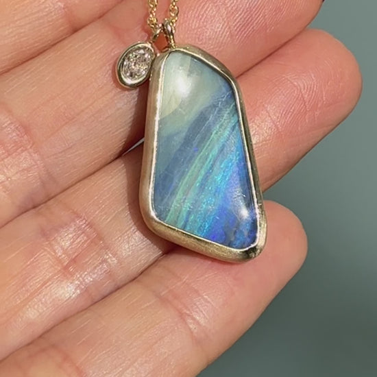 Video of an Australian Opal Necklace by NIXIN Jewelry showing the boulder opal pendant being gently turned to reveal the blue opal color shift and the textured ironstone back. This luxury jewelry video highlights the contrast between the polished diamond charm and the matte bezel setting, as well as the polished gold waves lapping the back of the opal stone.