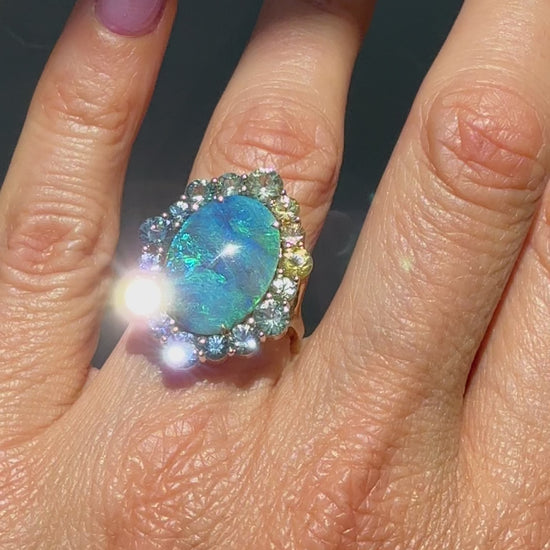 An Australian Opal Ring by NIXIN Jewelry. modeled on a hand. This video details the unique jewelry art of a modern opal ring. The luxurious craftsmanship highlights the black opal's brilliance and the vibrant colors of the ombré sapphire halo.