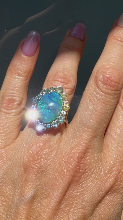 An Australian Opal Ring by NIXIN Jewelry. modeled on a hand. This video details the unique jewelry art of a modern opal ring. The luxurious craftsmanship highlights the black opal's brilliance and the vibrant colors of the ombré sapphire halo.