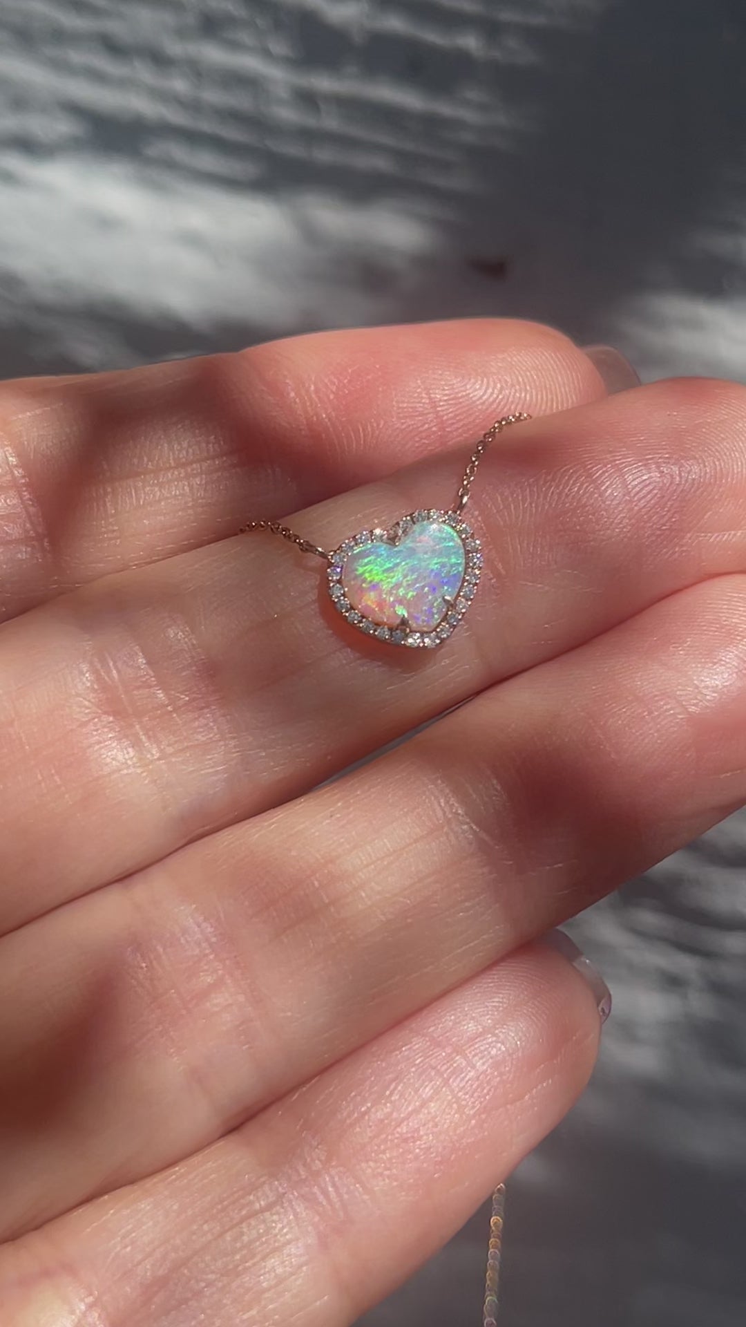 Video of an Opal Heart Necklace by NIXIN Jewelry moving in the sunlight. The pink and blue Crystal Opal flashes rainbow colors as the diamond halo sparkles around it.