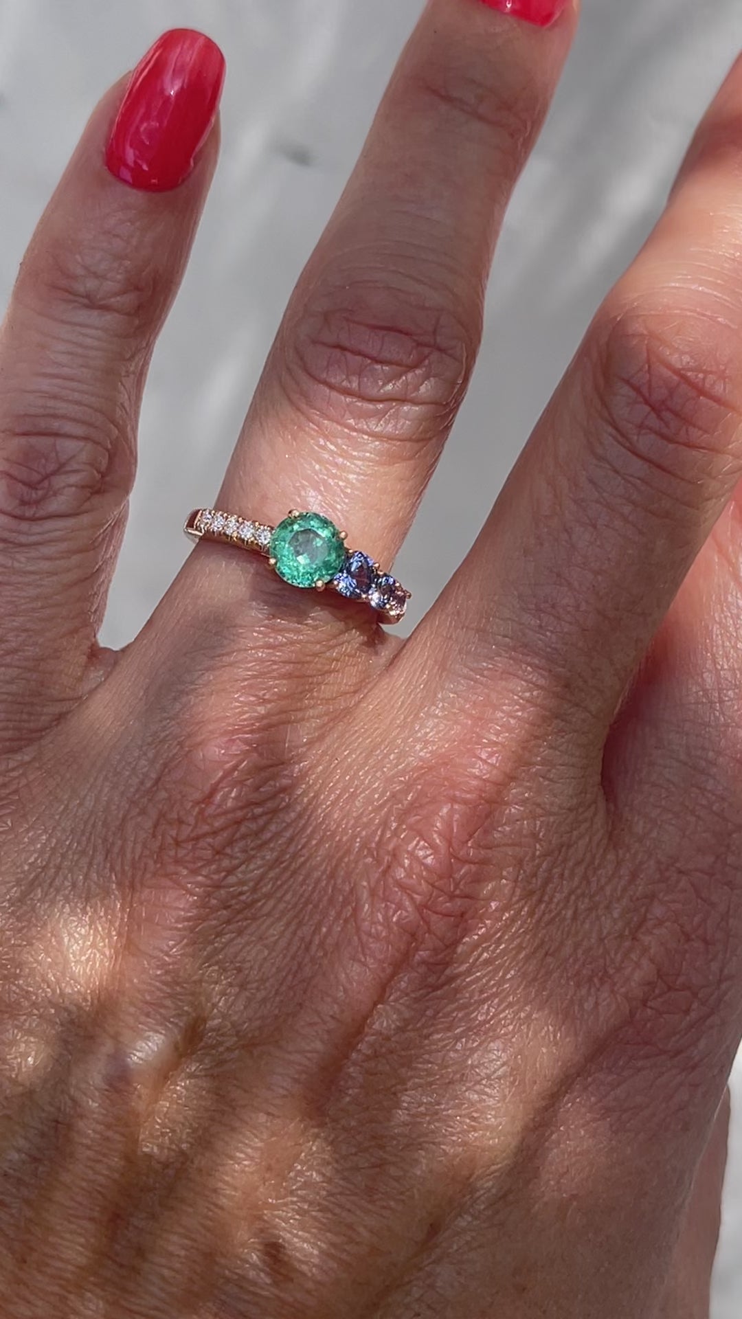 A Colombian Emerald Ring by NIXIN Jewelry modeled on the hand. The emerald diamond ring is made in rose gold and has purple sapphires cascading down one side.