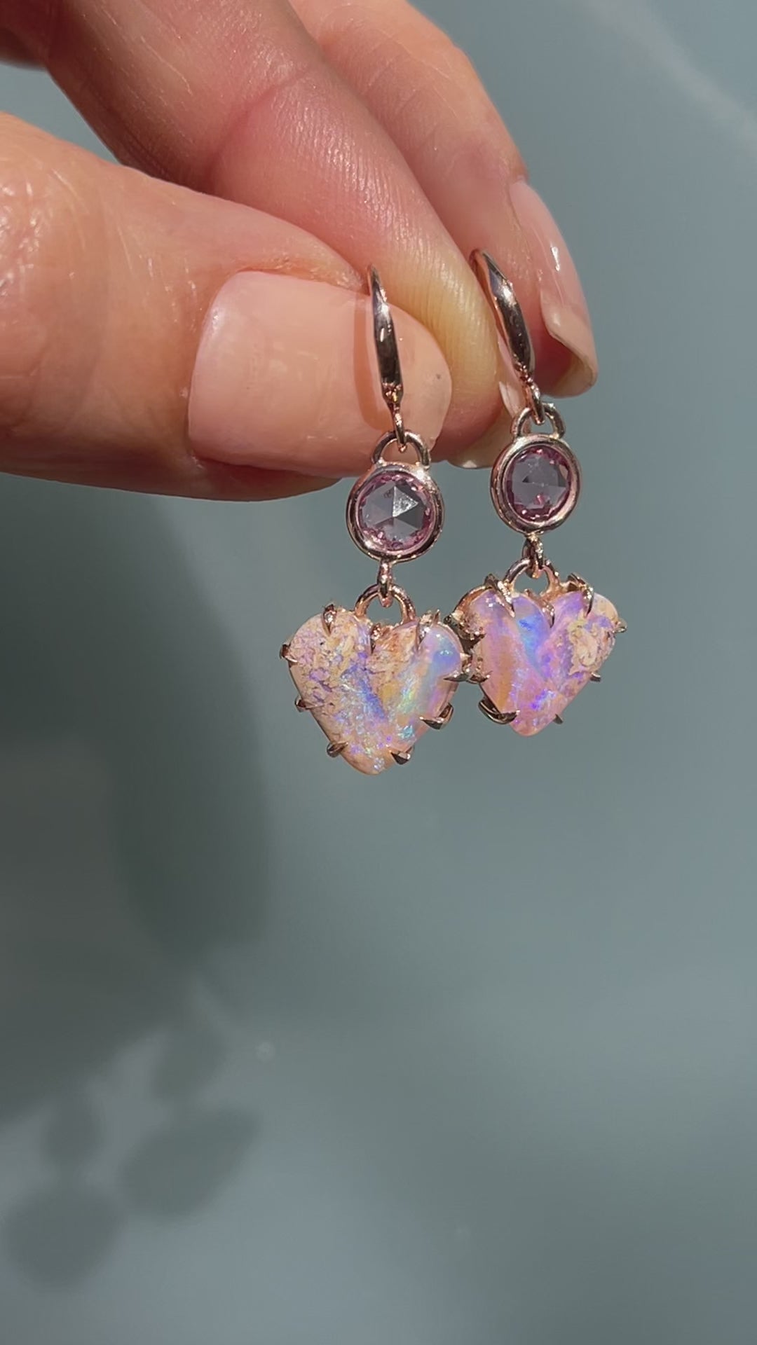 Video of a pair of Australian Opal Earrings by NIXIN Jewelry held in the sunlight. The heart earrings have purple opals and sapphires.