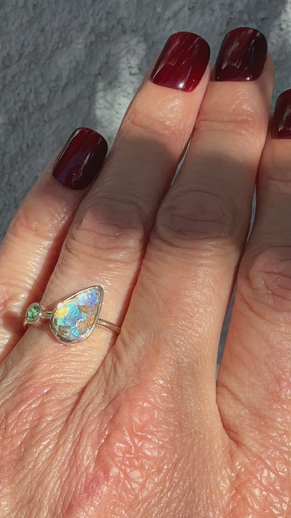 This Australian opal ring by NIXIN Jewelry is showcased in a video that captures the shifting play-of-color across its vibrant surface. As the hand moves, the pear shaped opal reveals deep flashes of lavender and electric blue, while the brilliant emerald flickers like a vivid green beacon. This high-definition look at the unique fine jewelry highlights the secure bezel setting and the modern design, making it an exquisite example of rare opal jewelry.