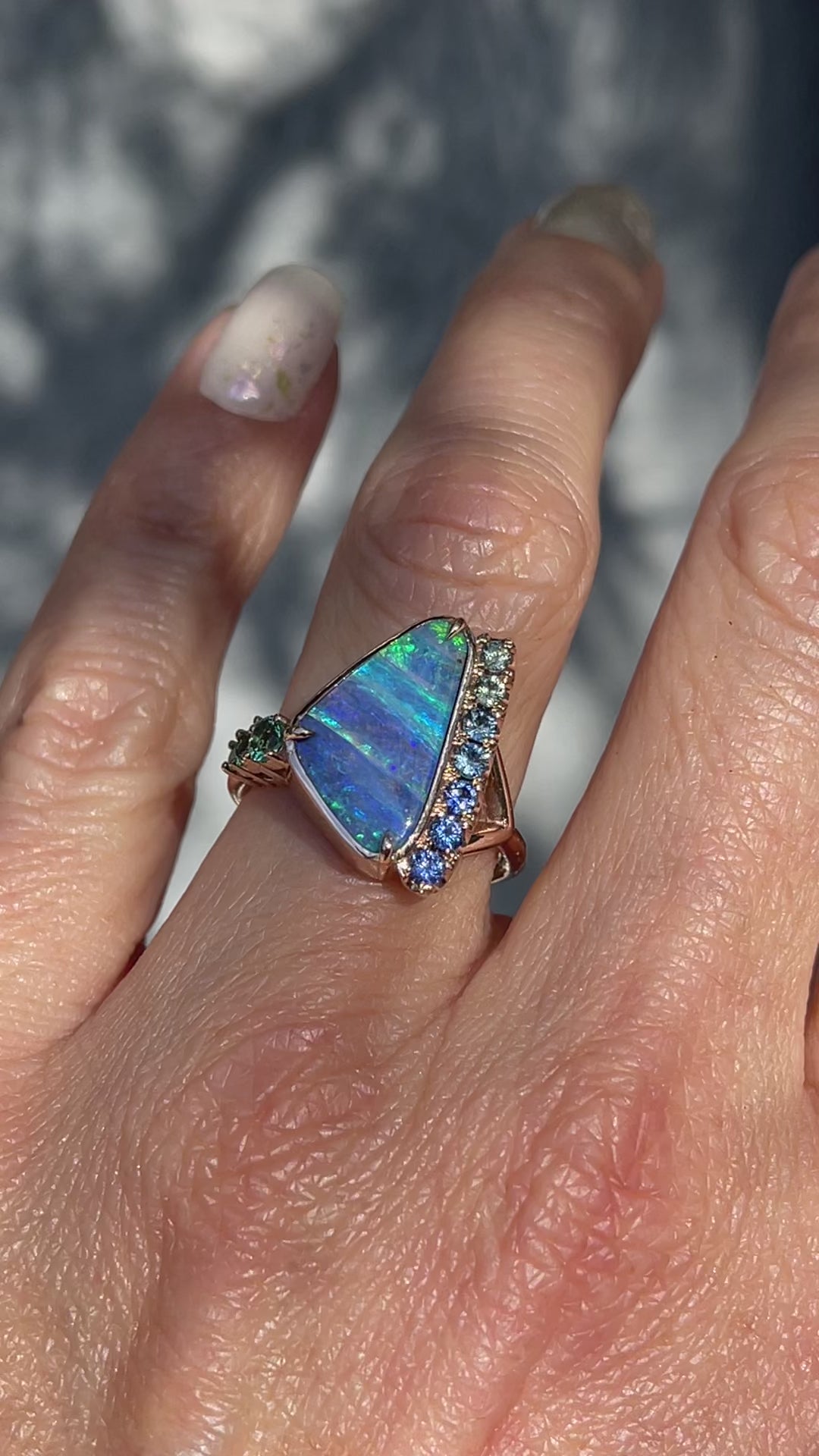 Video of an Australian Opal Ring by NIXIN Jewelry on a hand. The blue opal ring has sapphires and emeralds set in rose gold.