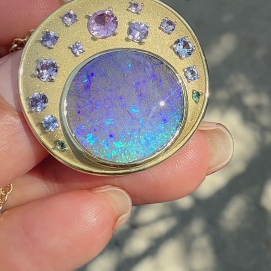This video displays an Australian Opal Pendant by NIXIN Jewelry, rotating to showcase a blue boulder opal in a bezel setting and a hidden tree engraving on the reverse. The luxury necklace features ombre sapphires in prong settings set against satin finish matte gold. This unique fine jewelry is a masterpiece of art, making this rare opal necklace a premier example of women’s luxury jewelry.