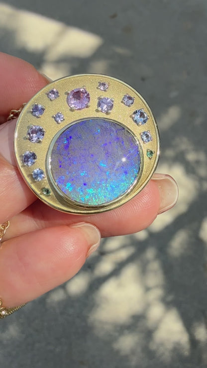 This video displays an Australian Opal Pendant by NIXIN Jewelry, rotating to showcase a blue boulder opal in a bezel setting and a hidden tree engraving on the reverse. The luxury necklace features ombre sapphires in prong settings set against satin finish matte gold. This unique fine jewelry is a masterpiece of art, making this rare opal necklace a premier example of women’s luxury jewelry.