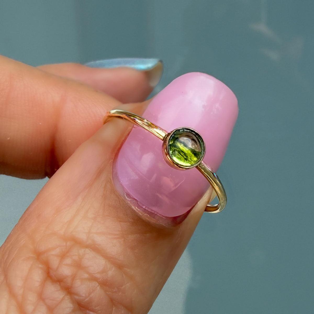 Lucky Green Tourmaline Ring held on a fingertip, a single soft olive tourmaline cabochon bezel set in 14k gold on a slim band.