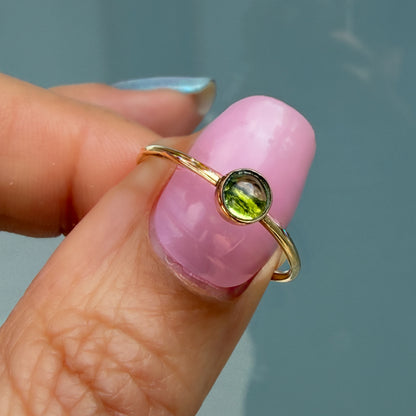 Lucky Green Tourmaline Ring held on a fingertip, a single soft olive tourmaline cabochon bezel set in 14k gold on a slim band.