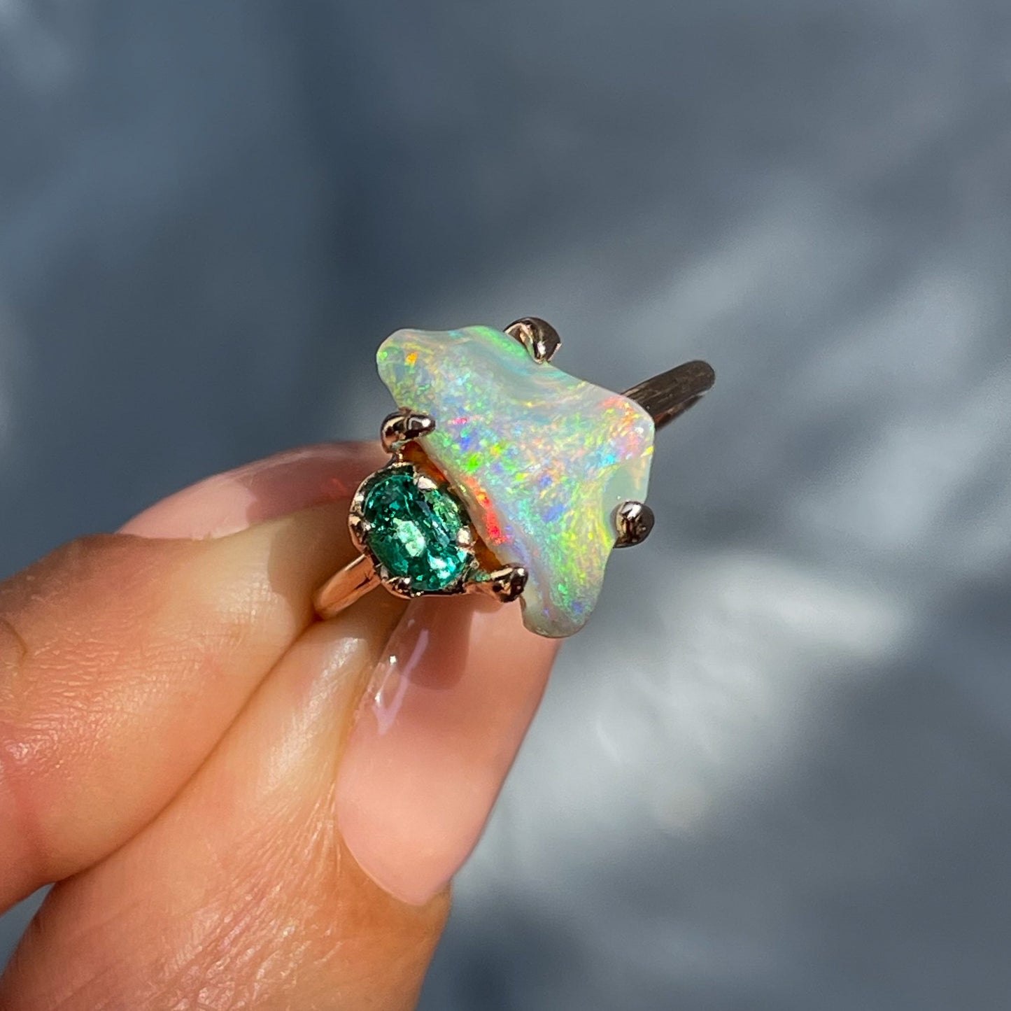 An Opal and Emerald Ring by NIXIN Jewelry held in front of a grey backdrop. The Australian Black Opal is set next to an oval emerald.