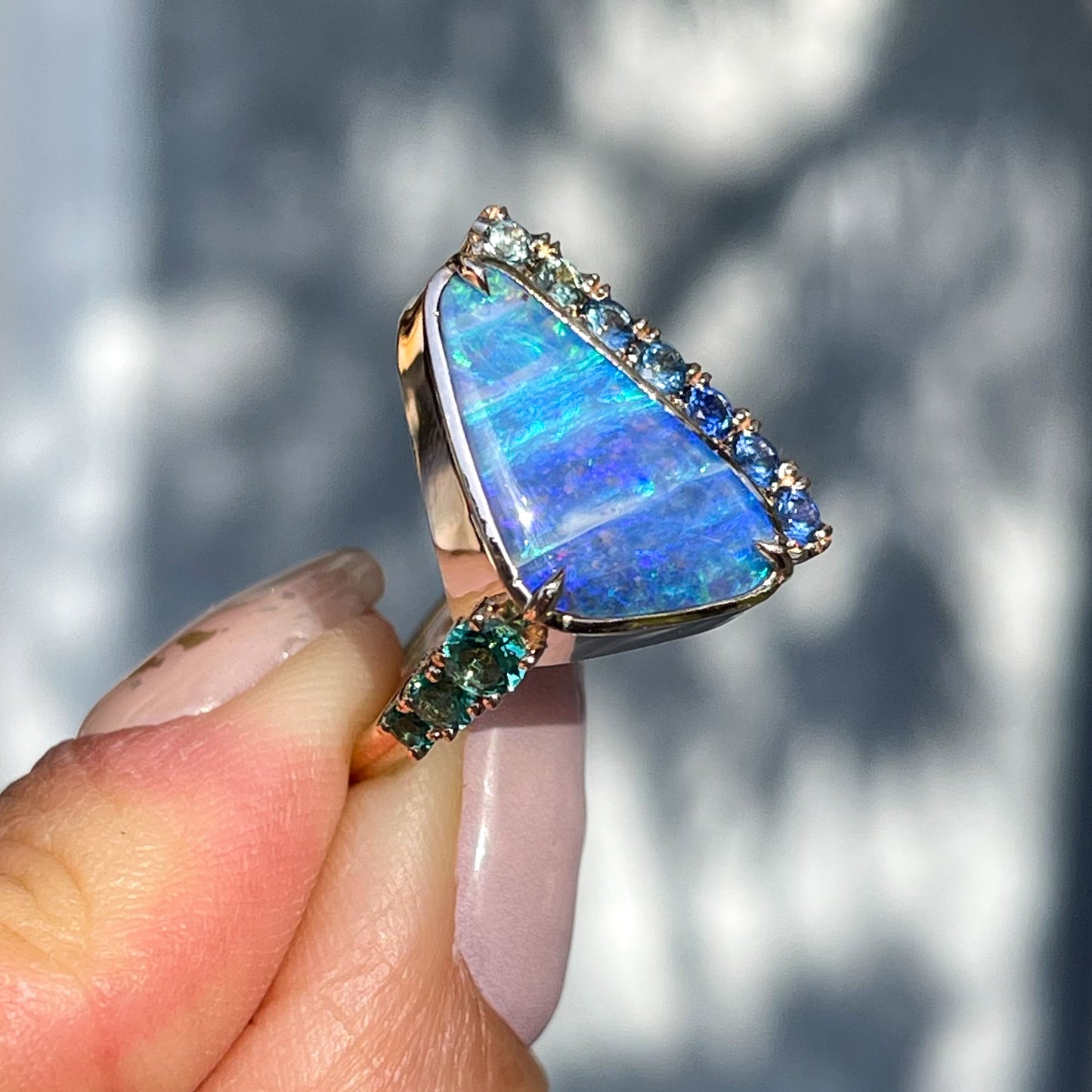 Angled view of an Australian Opal Ring by NIXIN Jewelry showing emeralds, sapphires and a blue opal in rose gold.