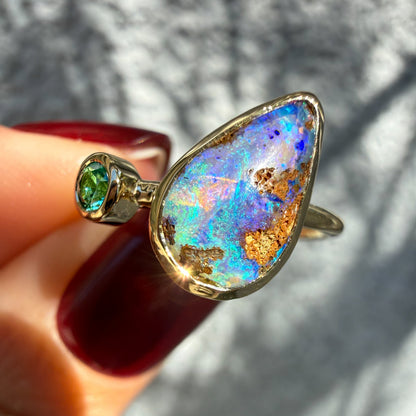 This close-up photograph provides a tilted perspective of an Australian Opal Ring by NIXIN Jewelry held between fingers with deep red nails. The view emphasizes the complex ironstone matrix embedded within the natural opal stone next to the vibrant green emerald. This dynamic pairing creates a piece of unique opal jewelry suitable as a cocktail ring.