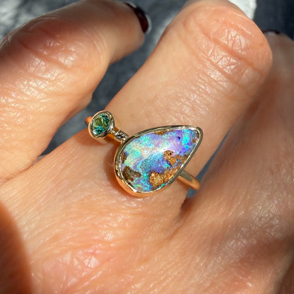 This is an Australian Opal Ring by NIXIN Jewelry. This perspective on the worn ring showcases the beautiful boulder opal jewelry characteristic of Australian stones, paired alongside a sparkling round emerald. This combination of materials makes for a piece of rare opal jewelry suitable for both a cocktail ring and daily wear.