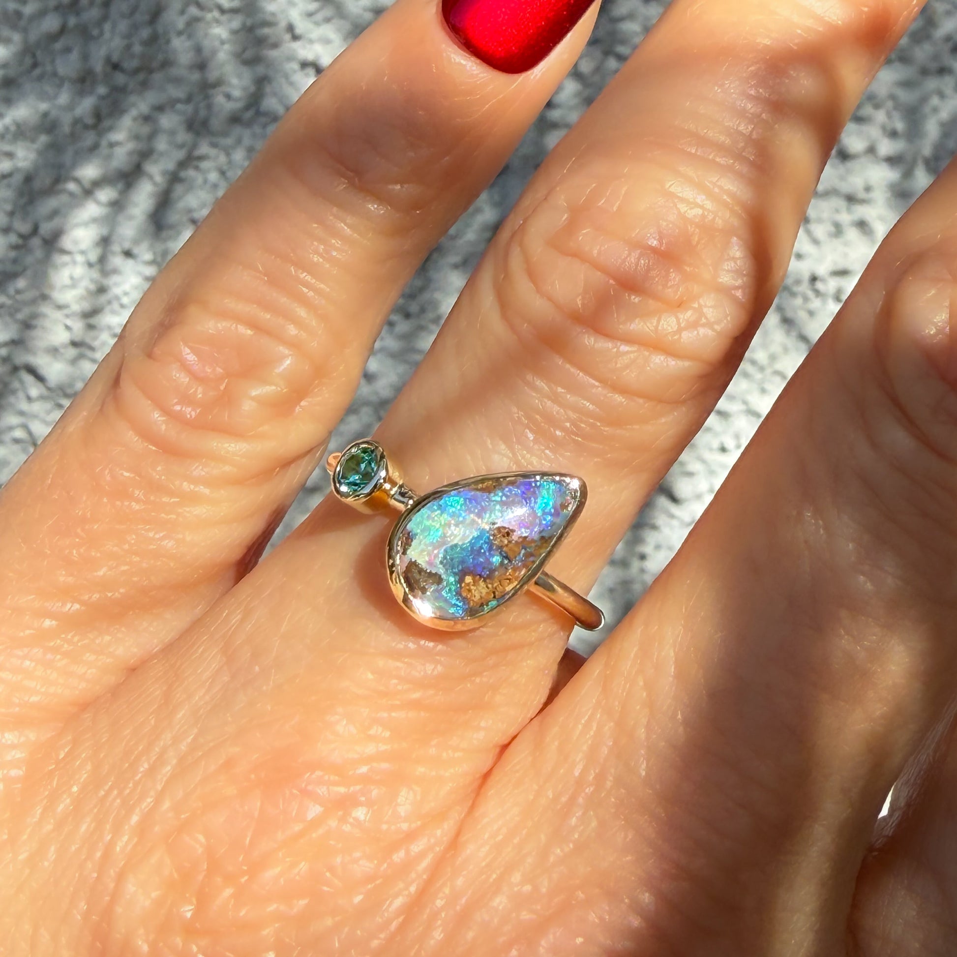 A detailed image showing an Australian Opal Ring by NIXIN Jewelry being worn on a hand with red fingernails under contrasting bright sunlight. The intense play-of-color within the rare opal ring highlights blues, purples, and greens next to a sparkling round emerald. This piece of wearable jewelry art is quintessential NIXIN.