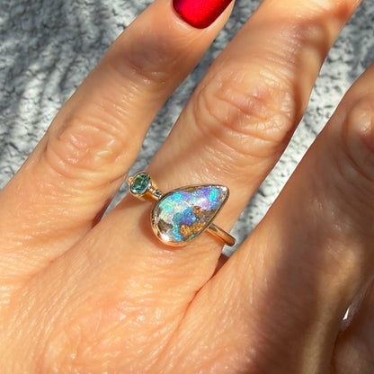 A detailed image showing an Australian Opal Ring by NIXIN Jewelry being worn on a hand with red fingernails under contrasting bright sunlight. The intense play-of-color within the rare opal ring highlights blues, purples, and greens next to a sparkling round emerald. This piece of wearable jewelry art is quintessential NIXIN.