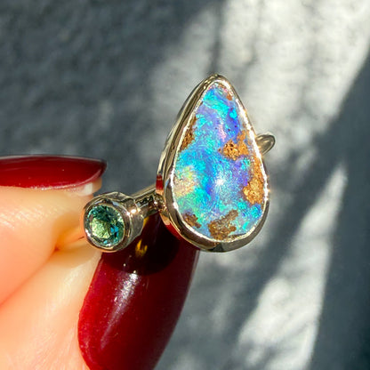 This is an Australian Opal Ring by NIXIN Jewelry. It is a unique luxury ring handcrafted in yellow gold opal ring featuring a pear-shaped natural opal and a round emerald, both in classic bezel setting. This modern design makes a stunning statement as a unique piece of fine jewelry art.