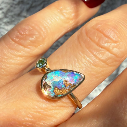 This is an Australian Opal Ring by NIXIN Jewelry being worn. The vibrant blue opal ring displays intense play-of-color, catching the light alongside a sparkling faceted emerald. This bold cocktail ring style exemplifies rare opal jewelry, perfect for those seeking unique fine jewelry.