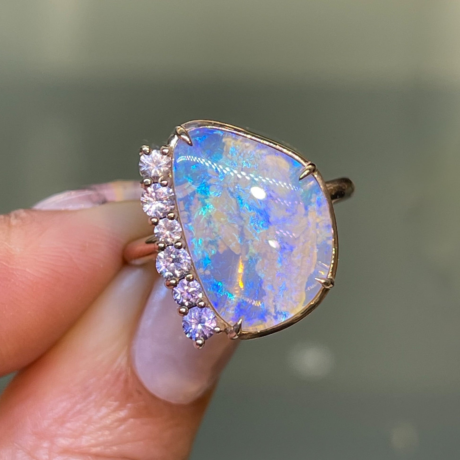 An Australian Opal Ring by NIXIN Jewelry held up under indoor lighting. The opal and sapphire ring is set in 14k rose gold.
