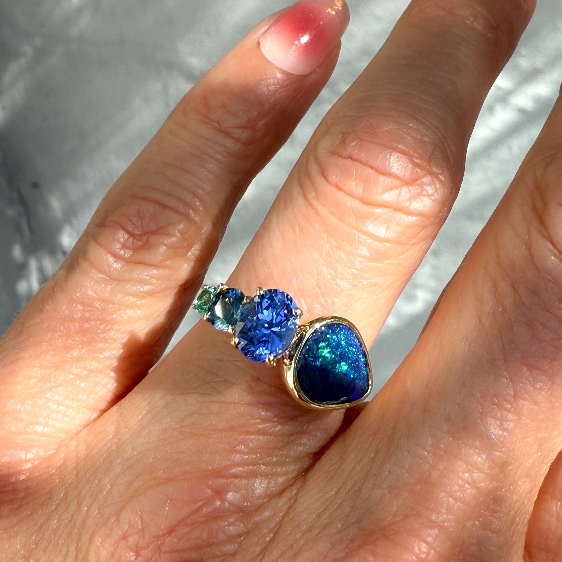 An Australian Opal Ring by NIXIN Jewelry. This vibrant image of the gold opal ring worn on a hand highlights the dual pattern within the australian opal and the stunning blue tones of the sapphire and emerald side stones.