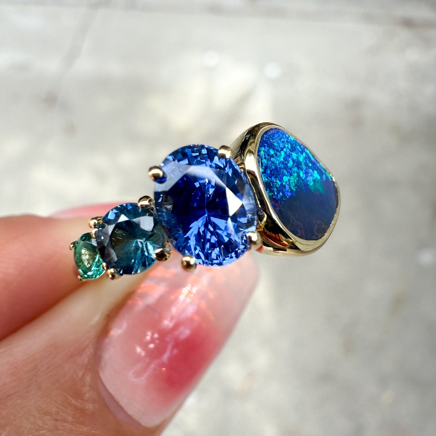 An Australian Opal Ring by NIXIN Jewelry. A close-up view of the ring shows the beautiful contrast between the black opal and the saturated blue and teal sapphires and emerald in a modern opal ring design.